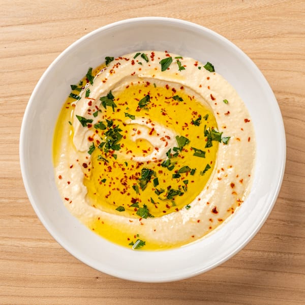 Nour's Lebanese hummus, drizzled with olive oil, parsley, and red pepper flakes in a white bowl.