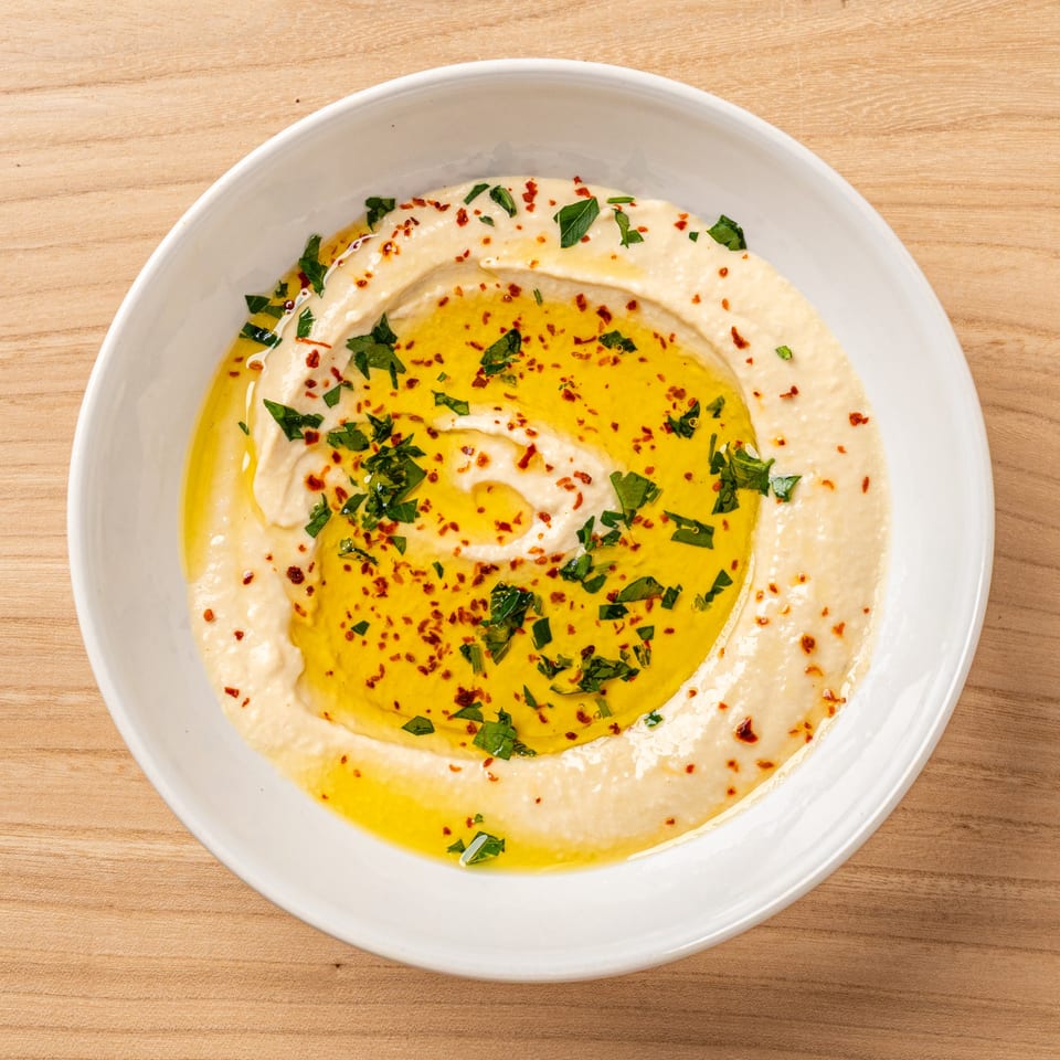Nour's Lebanese hummus, drizzled with olive oil, parsley, and red pepper flakes in a white bowl.