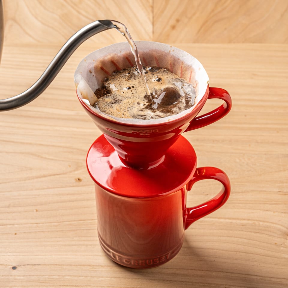 Pour-over coffee being brewed into a red mug on a light wooden surface.